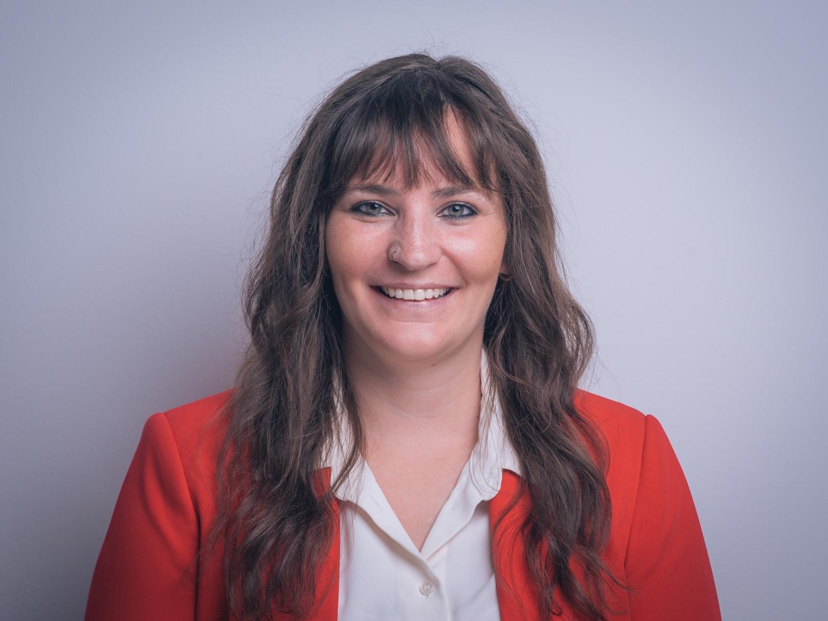 Portr&auml;t einer l&auml;chelnden Frau mit langen, lockigen Haaren, in einem roten Blazer und wei&szlig;em Hemd, vor einfarbigem Hintergrund.