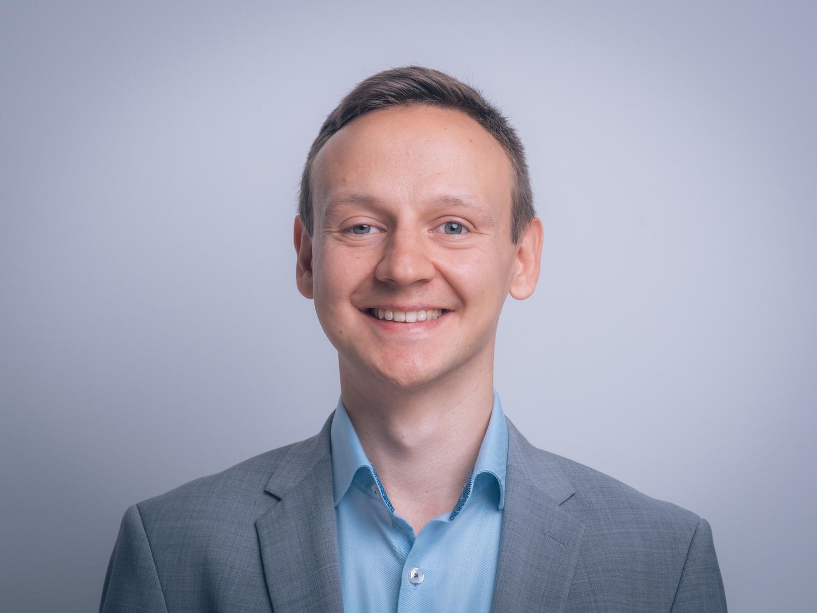 Portr&auml;t eines l&auml;chelnden Mannes in grauem Anzug und hellblauem Hemd vor einem hellen Hintergrund.